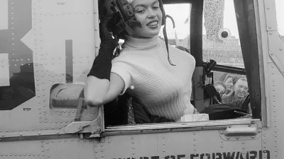 Black and white photo of actress Jayne Mansfield leaning out of the window of a military plane wearing flight helmet for promotional photo