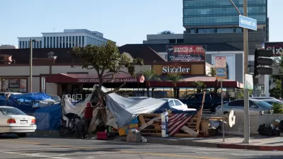 A homeless encampment fills a sidewalk in front of a Sizzler restaurant advertising Thanksgiving dinner.