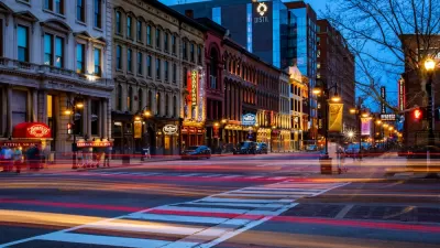 A Downtown Louisville street at night, with the lights of passing shown with a blurred effect.