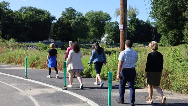 Group of people walking on paved walkway in Naugatuck River Greenway on sunny day next to green field.