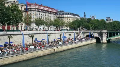 A large crowd gathers on the banks of the River Seine on a sunny day in Paris.