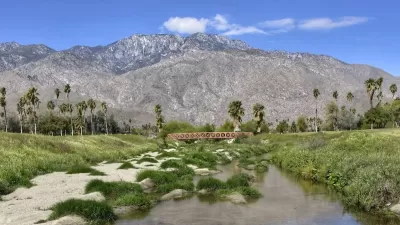 View of small pedestrian bridge in Prescott Preserve Palm Springs, California with palm tree oasis and mountains in background.