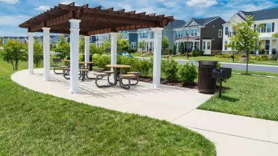 A pergola with a grill is located in a grassy park, across the street from single-family housing in a suburban style.