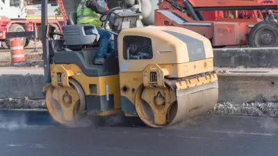 A man operates a yellow roller to smooth out fresh pavement on a road.