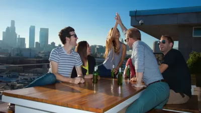 A group of friends laughs and celebrates with beers on a rooftop with Downtown Los Angeles in the background.