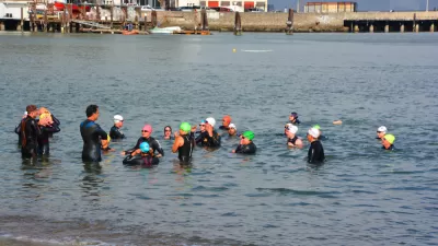 A group of wetsuit-clad swimmers gathers to talk in shallow water near the shore of the San Francisco Bay.
