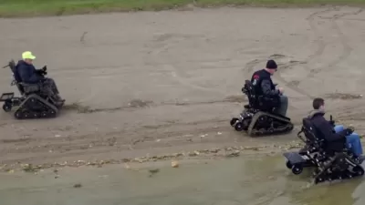 Aerial view of three men using track chair mobility devices on lake beach in Michigan state park.