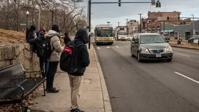 People in hoodies and jackets waiting for oncoming bus at sidewalk bus stop in Boston, Massachusetts.