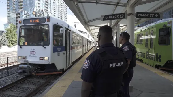 Two transit police officers standing on outdoor train platform in Denver with white light rail train approaching.