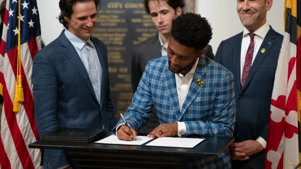 Mayor Brandon M. Scott of Baltimore wearing blue checkered suit signing a bill with three men in suits standing behind him at podium.