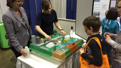 Two adults stand behind a table, gesturing toward a model of a city while talking to a young child about buildings and parks and streets.