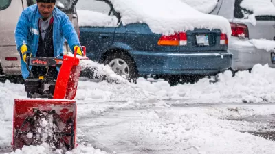 A man pushes a snow blower to clear the ground of an outdoor parking lot filled with cars.