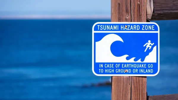 A sign, with the blue ocean int he background, reads "Tsunami Hazard Zone: In Case of Earthquake Go to Higher Ground or Inland"