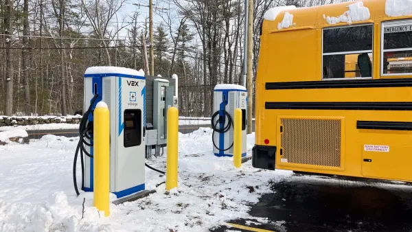 Yellow electric school bus plugged into vehicle-to-grid charging station in snowy parking lot.