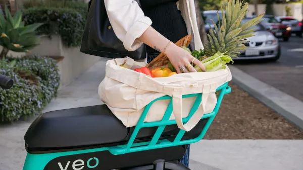 Close-up on back basket of teal and black Veo bike share bike with woman's hands reaching into canvas bag full of produce.