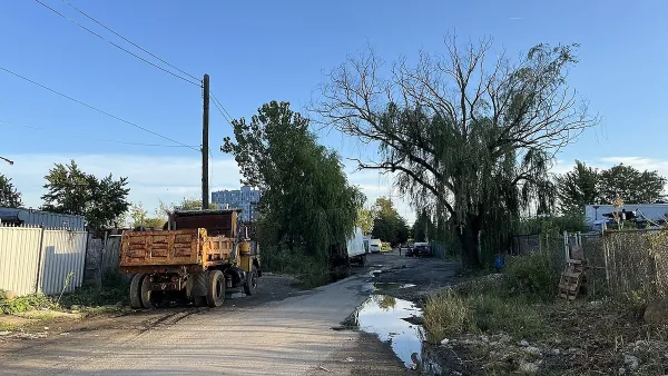 Small paved street with yellow dump truck parked on side and puddle on shoulder in Brooklyn.