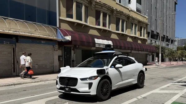 White Waymo robotaxi on street in Miami, Florida with two men walking on sidewalk in background.