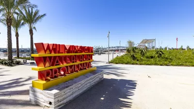 Person-height red 'Wilmington Waterfront Promenade' sign on concrete walkway with palm trees in background on waterfront walkway at the Port of Los Angeles, California.