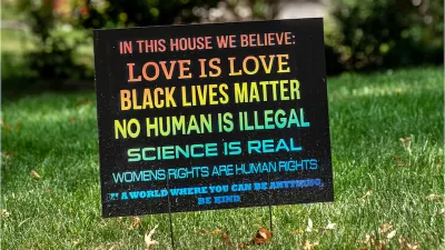 A rainbow colored sign planted in the front yard of a typical suburban home reads, "In this house we believe love is love, black lives matter, no human is illegal, science is real, women's rights are human rights. In a world where you can be anything, be kind."