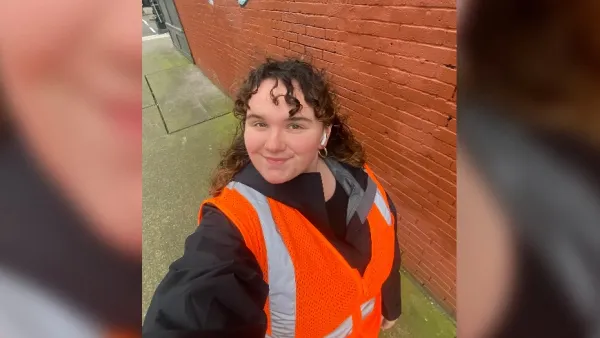 Woman taking selfie while wearing orange safety vest outdoors in front of red brick wall on field visit.