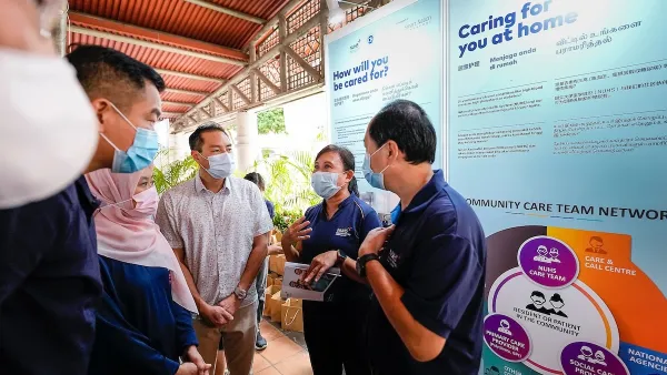 Two people in masks stand in front of a "caring for you at home" sign. They speak to people in medical masks.