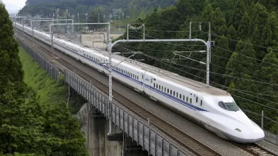 Shinkansen high-speed rail train traveling across bridge with green trees on either side.