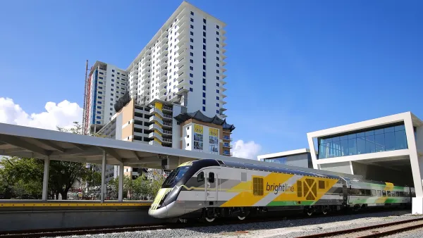 Brightline train driving out from station with tall apartment building in background.