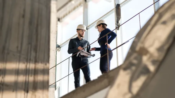 Man and woman in white hard hats standing on balcony at job site.