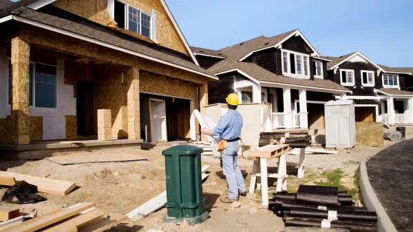 Man in yellow hard hat holding plan and looking up at single-family homes under construction.