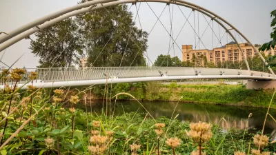 View of pedestrian suspension bridge over waterway surrounded by green grass with small apartment buildings in background
