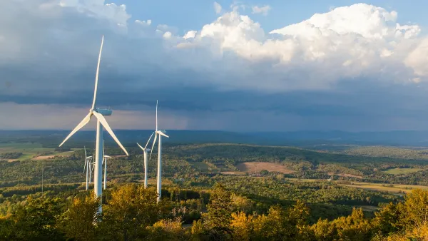 A cluster of wind turbines beneath a stormy sky, above a forest where the leaves are beginning to change colors.