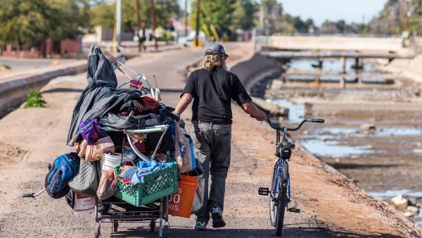 Unhoused man pushed a shopping cart filled with items and a bicycle along an empty canal in Phoenix, Arizona.
