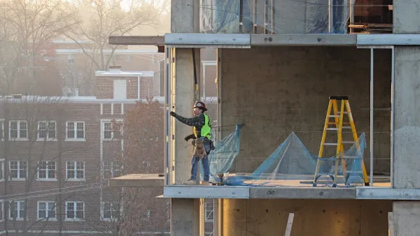 Construction worker in safety gear stands in an unfinished concrete high-rise building with older brick buildings visible in background.