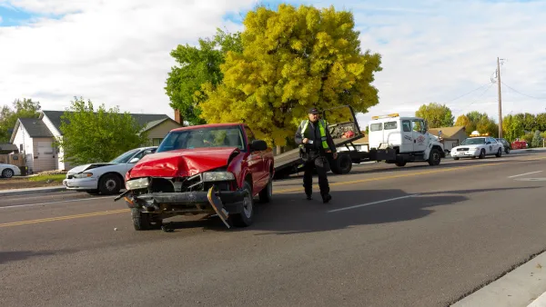 Tow truck driver stands next to red sedan with crumpled front in middle of street.