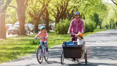 Father riding cargo bike with one child in cargo basket and one child riding bike alongside on sunny tree-lined path