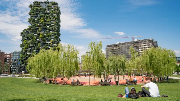 People sitting on grass in park in Milan, Italy with grove of willows and a vertical forest skyscraper in background.
