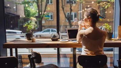 Woman sitting at cafe table with laptop, holding coffee cup and looking out at street
