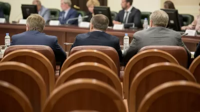 View from back of three men in suits sitting in a large room with leather chairs facing dais