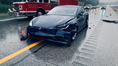 Dark blue Tesla with crumpled front bumper on rainy highway with red fire truck in background