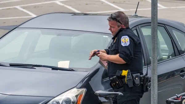 Male parking enforcement officer in uniform stands next to car with ticket in its windshield talking on phone or radio.