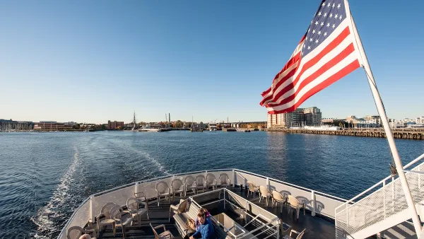 View from on board a Boston ferry with people sitting in front and an American flag flying from the boat on a sunny day.