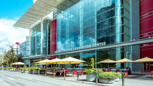 Front of George R. Brown convention center in Houston, Texas with yellow umbrellas over red tables in front.