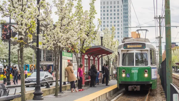 Green MBTA tram pulling up to station with people waiting under a flowering tree in Boston, Massachusetts.