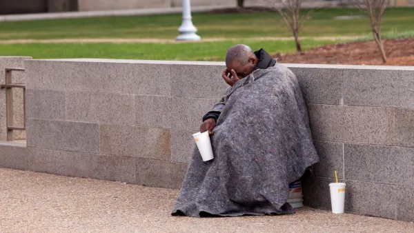 Unhosued man wrapped in grey blanket crouches next to concrete wall holding out white paper cup for change.