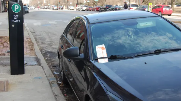 Dark-colored car parked at curb with white and red parking ticket in passenger side windshield wiper next to parking pay station with green P sign on sidewalk.