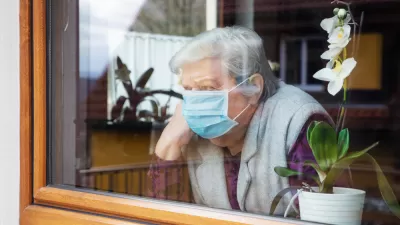 Elderly woman with short gray hair wearing blue surgical mask looks out window with wooden frame, a potted purple orchid on the windowsill