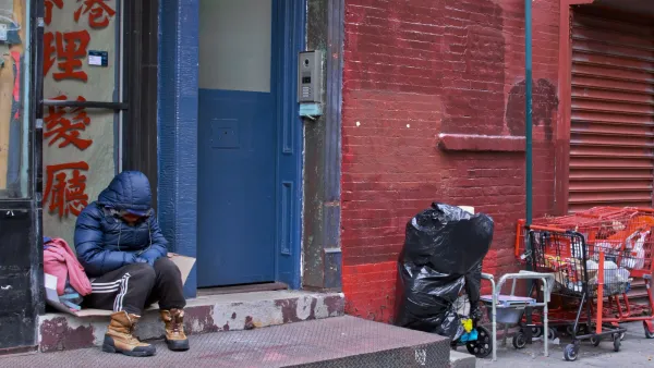 Unhoused person in blue parka sits huddled on a chair against a blue exterior door on red brick wall in New York City.