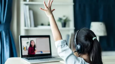 View from behind young girl student wearing headphones and raising her hand as she attends online class with dark-haired female teacher on laptop in front of her