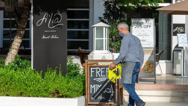 Man using lime green leaf blower in front of restaurant with sandwich board advertising mimosas in Miami Beach, Florida.