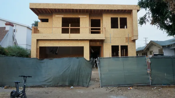 Boxy wood-frame two-story house under construction behind green construction fence in Los Angeles, California.
